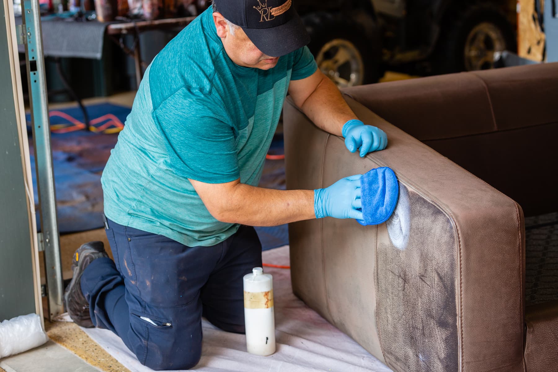 A person wearing blue gloves wipes a white product onto the side of a brown leather chair